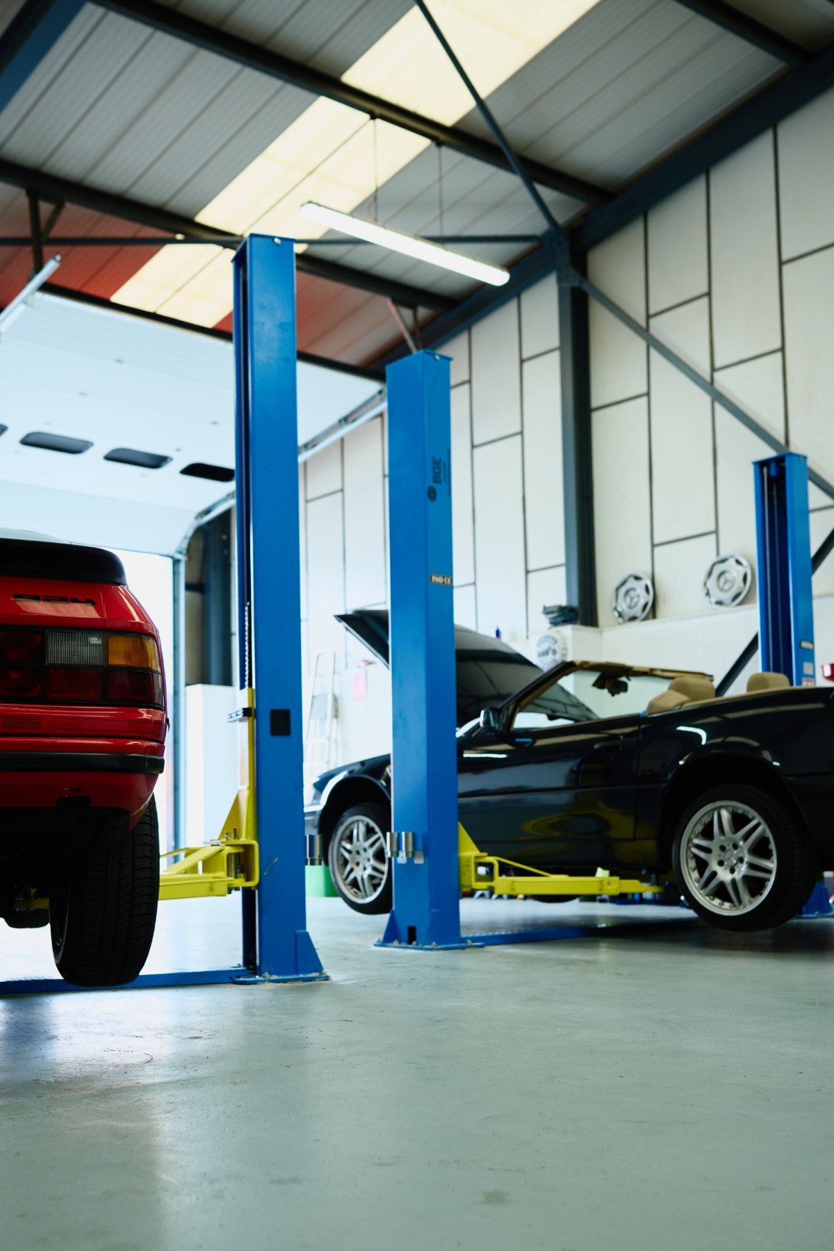 A red car and a black convertible on two-post lifts in a bright workshop